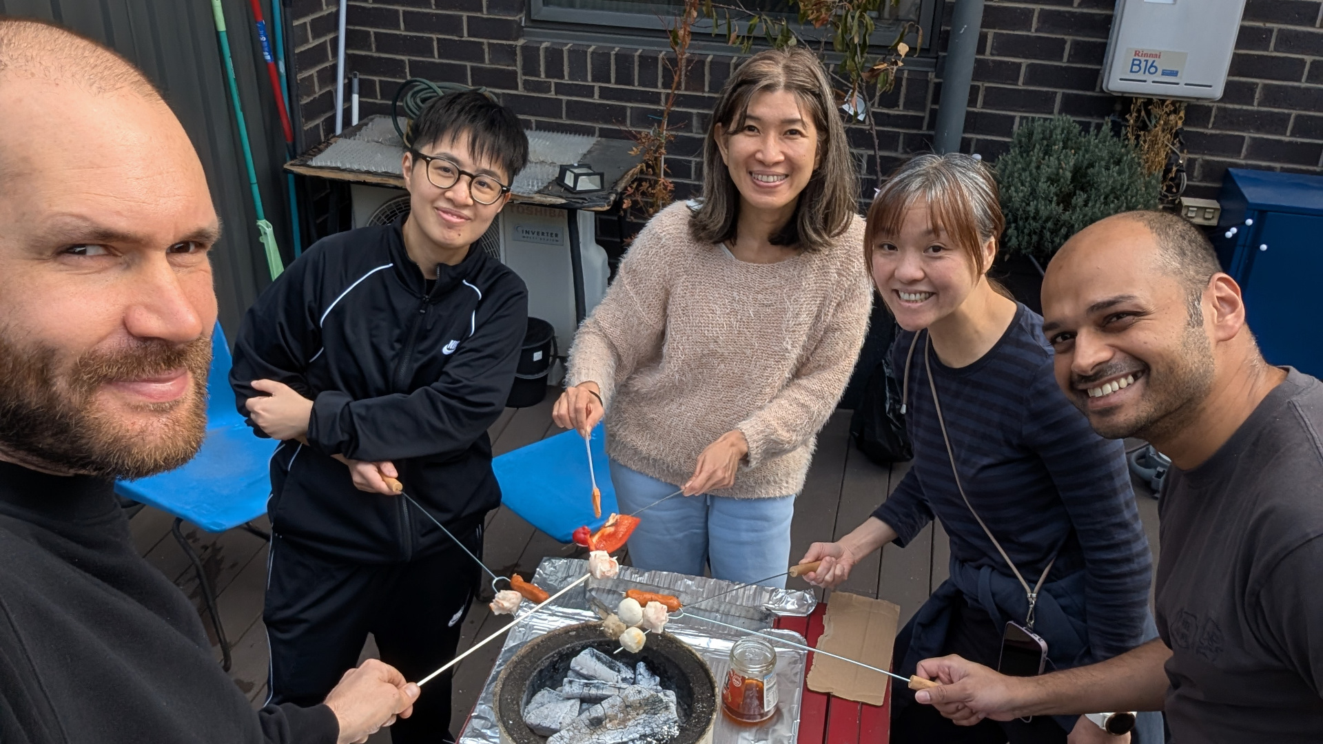 A selfie of five people outdoors on a deck, holding skewers with food over a clay-pot fireplace.